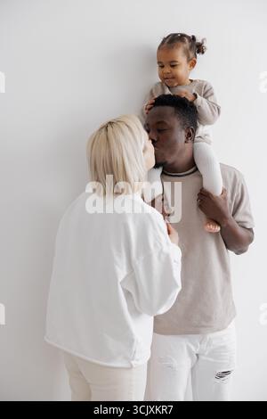 Famille biraciale aimante avec leur fille assise sur les épaules du père, partageant un moment tendre contre un mur blanc. Concept de famil interracial Banque D'Images