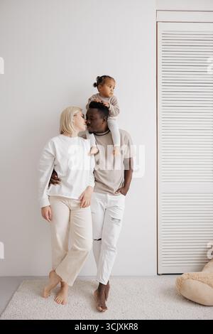 Famille biraciale aimante avec leur fille assise sur les épaules du père, partageant un moment tendre contre un mur blanc. Concept de famil interracial Banque D'Images