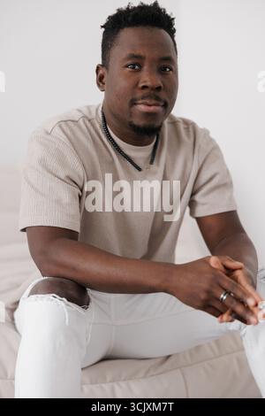 Élégant homme africain dans un t-shirt beige et Jean blanc déchiré s'assoit et regarde directement l'appareil photo. Banque D'Images