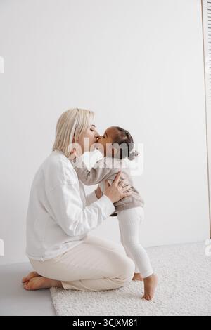 Mère aimante s'agenouille et partage un baiser tendre avec sa fille biraciale dans une pièce lumineuse et simple. Concept de famille interraciale. Banque D'Images