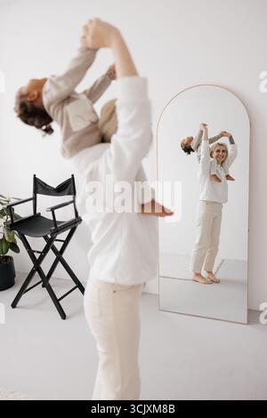 Happy Mother élève sa fille biraciale haut dans les airs, capturée dans un reflet miroir dans une pièce lumineuse et minimaliste. Concept de famille interraciale. Banque D'Images
