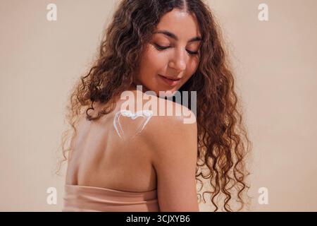 Le modèle féminin avec des cheveux bouclés affiche la conception de coeur sur l'épaule, posant sur fond beige doux, soulignant la beauté, la confiance et l'expré artistique Banque D'Images