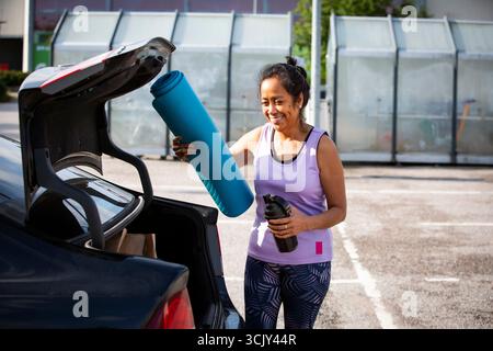 La femme d'âge moyen confiante de couleur prend tapis de yoga du coffre de voiture, montrant l'équilibre de la vie urbaine, la diversité et le mode de vie sain. Banque D'Images