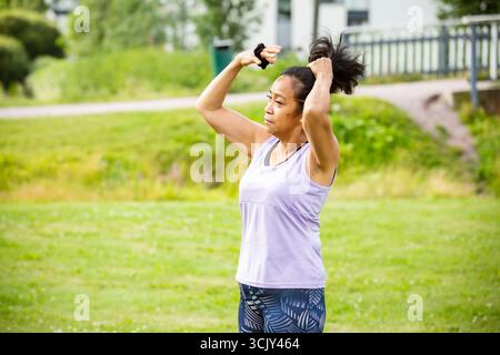 Femme d'âge moyen confiante de couleur liant les cheveux pour le yoga en plein air dans le parc, favorisant le bien-être, la pleine conscience, la forme physique et un style de vie actif. Banque D'Images