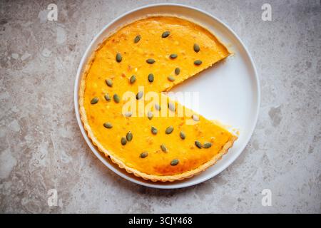Tarte à la citrouille avec une tranche découpée, surmontée de graines de citrouille, présentée sur une assiette blanche, soulignant la texture du dessert et son aspect invitant Banque D'Images