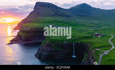 Découvrez la vue imprenable sur la cascade Mulafossur qui se déverse sur les falaises près du village de Gasadalur dans les superbes îles Féroé, illuminée par la lueur chaude d'un coucher de soleil. Banque D'Images
