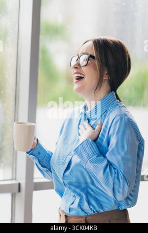 Jeune femme d'affaires riant avec une tasse à café dans un cadre de bureau moderne lumineux profitant d'un moment joyeux Banque D'Images