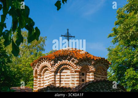 Dôme d'église orthodoxe traditionnel avec tuiles de toit en terre cuite rouge et croix décorative en métal, encadrée par des arbres verts et un ciel bleu clair par un été ensoleillé Banque D'Images