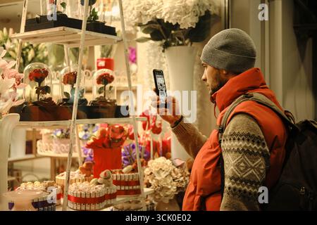 Homme en gilet rouge photographie des décorations de magasin de fleurs à travers la fenêtre en verre la nuit. Banque D'Images