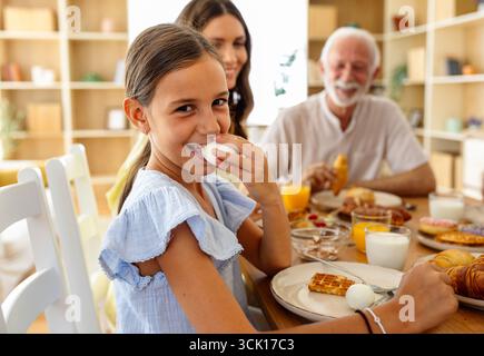 Une fille mange un œuf tandis que d'autres à la table sourient. De délicieux petits déjeuners les entourent dans une salle à manger confortable. Banque D'Images