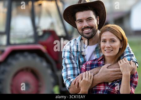 Couple fermier souriant debout tôt le matin dans le champ avec tracteur en arrière-plan. Homme embrassant sa jolie petite amie par derrière Banque D'Images