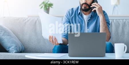 Homme d'affaires arabe irrité travaillant à la maison, assis sur un canapé avec un ordinateur portable, tenant des documents et ayant un appel téléphonique Banque D'Images