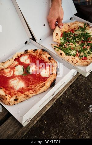 Deux pizzas gourmandes dans des boîtes ouvertes placées sur un banc de parc en bois, avec la main masculine saisissant une tranche, serrée contre des pelouses vertes et des parterres de fleurs, Outdoo décontracté Banque D'Images