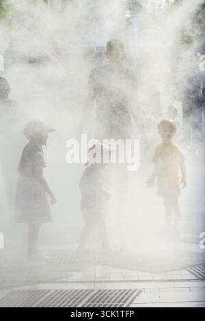 Enfants marchant dans la brume ou l'eau pulvérisée par une journée chaude. La scène capture la joie et le soulagement de se rafraîchir pendant l'été Banque D'Images