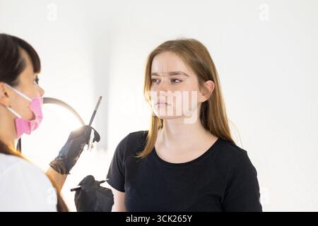 Une femme portant une chemise noire se fait façonner ses sourcils par un professionnel de la beauté. Le professionnel porte un masque facial rose et des gants noirs A. Banque D'Images