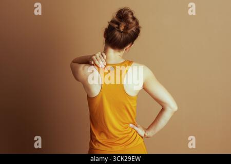 Une femme en dos de nageur orange et leggings assortis se tient avec le dos à la caméra, massant l'épaule. Cette image capture un moment de malaise physique Banque D'Images