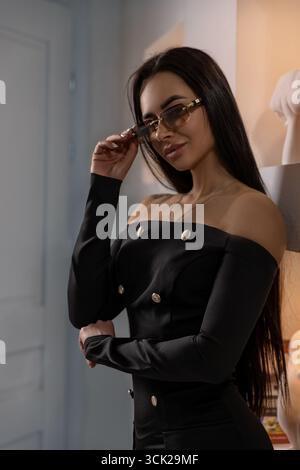 fille avec des lunettes montre la confiance comme elle pose soigneusement à l'intérieur. Elle porte une tenue noire élégante avec des boutons dorés mettant en valeur son sens de la mode. Banque D'Images