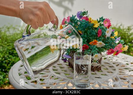 Une photo d'action d'une personne versant de l'eau potable propre dans un cadre de jardin vibrant avec des fleurs colorées. Un concept de rafraîchissement, d'hydratation, un hea Banque D'Images