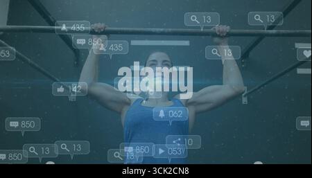 Tirant vers le haut athlète féminin portant un haut athlétique bleu sur la barre de traction dans la salle de gym, avec des icônes de notification Banque D'Images