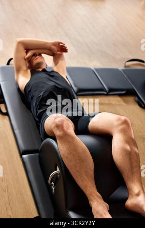 Un homme vêtu de vêtements de sport repose sur un appareil pilates, paraissant détendu après l'exercice. Le studio de remise en forme dispose d'un parquet et professionnel Banque D'Images