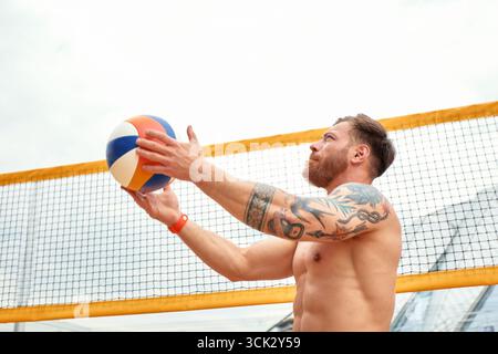 Un homme musclé avec des tatouages est vu jouer au volley-ball sur la plage. Il est engagé dans le jeu, mis contre un ciel lumineux et un court de sable, créant un Li Banque D'Images