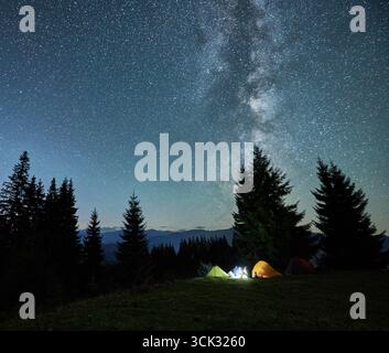Ciel étoilé nocturne au-dessus d'une colline herbeuse avec des tentes touristiques illuminées et des conifères. Vallée de montagne avec des tentes de camp sous le ciel avec des étoiles et la voie lactée. Randonnée, camping de nuit et concept d'astronomie. Banque D'Images