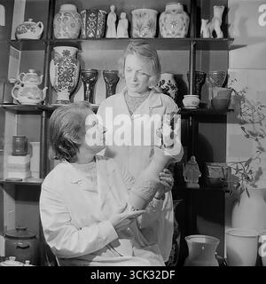Deux femmes artistes ou technologues dans le laboratoire d'art du Sloviansk Ceramic combine (Sloviansk, RSS d'Ukraine) discutent d'un nouveau produit dans les années 1980 La photographie d'archive capture un moment de travail où des spécialistes des manteaux blancs comparent une figurine émaillée finie à un vase non cuit. Les étagères remplies d’échantillons de produits finis soulignent l’atmosphère créative et la gamme de biens de consommation (TNP) produits par l’usine Banque D'Images