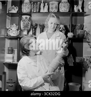 Deux femmes artistes ou technologues dans le laboratoire d'art du Sloviansk Ceramic combine (Sloviansk, RSS d'Ukraine) discutent d'un nouveau produit dans les années 1980 La photographie d'archive capture un moment de travail où des spécialistes des manteaux blancs comparent une figurine émaillée finie à un vase non cuit. Les étagères remplies d’échantillons de produits finis soulignent l’atmosphère créative et la gamme de biens de consommation (TNP) produits par l’usine Banque D'Images