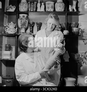 Deux femmes artistes ou technologues dans le laboratoire d'art du Sloviansk Ceramic combine (Sloviansk, RSS d'Ukraine) discutent d'un nouveau produit dans les années 1980 La photographie d'archive capture un moment de travail où des spécialistes des manteaux blancs comparent une figurine émaillée finie à un vase non cuit. Les étagères remplies d’échantillons de produits finis soulignent l’atmosphère créative et la gamme de biens de consommation (TNP) produits par l’usine Banque D'Images