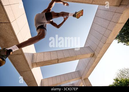 Prise de vue dynamique en bas angle d'un athlète sautant entre des structures architecturales contre un ciel clair et lumineux. La scène capture la force, l'équilibre et le Mo Banque D'Images