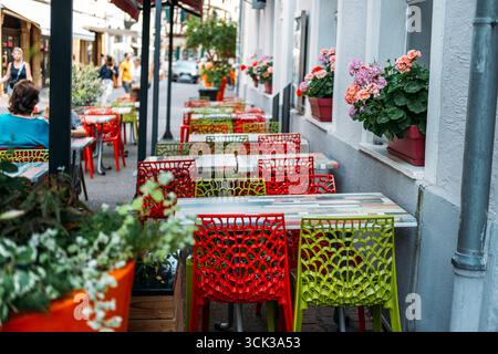 Terrasse de café colorée avec chaises en treillis rouge et vert bordant une rue européenne étroite ornée de boîtes à fleurs en géranium. Dynamisme urbain, café Banque D'Images