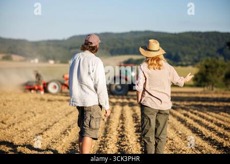 Deux agriculteurs regardent tracteur avec semoir semant la récolte dans les champs et la culture agricole. Travail d'équipe dans une ferme biologique Banque D'Images