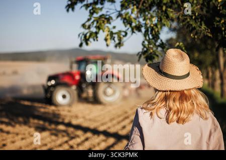 Femme fermière avec chapeau de paille regardant le tracteur avec semoir travaillant et semant la récolte sur le champ agricole Banque D'Images