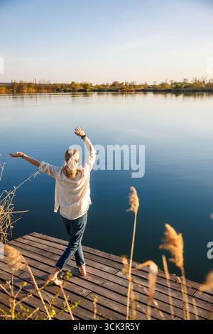 Femme insouciante debout sur la jetée au lac exerçant et dansant avec les bras levés dans la pose joyeuse, embrassant l'atmosphère tranquille de l'été indien Banque D'Images
