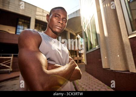 Un homme ajusté et musclé portant des vêtements décontractés se tient en toute confiance à l'extérieur. L'environnement urbain derrière lui est illuminé par la lumière du soleil. Sa détermination Banque D'Images