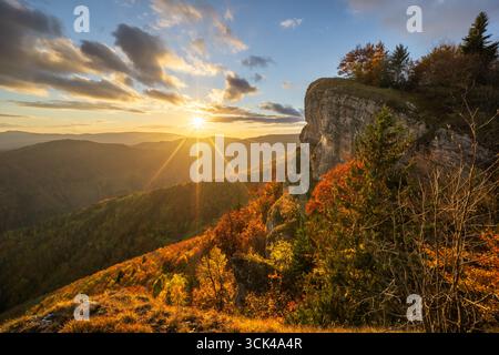 Vue du soleil doré se couchant derrière des sommets lointains, projetant une lueur chaude sur le feuillage d'automne accroché aux falaises rocheuses, rocher Majerova skala, Staré Hory, Banskobystrický kraj, Slovaquie. Banque D'Images
