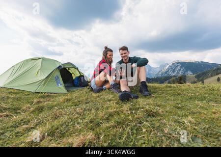 Heureux jeune couple campant, regardant quelque chose sur une tablette à l'extérieur de leur tente, partageant un moment joyeux entouré par la nature. Banque D'Images