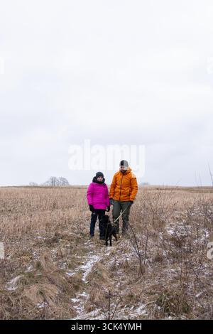 Deux individus se promènent dans un vaste champ herbeux avec leur chien noir, entouré d'une atmosphère hivernale sereine et d'un ciel doux et couvert. Banque D'Images