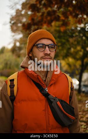 Homme avec des lunettes en tenue d'automne marche sur un chemin de parc couvert de feuilles, portant un gilet rouge et un bonnet orange. Banque D'Images