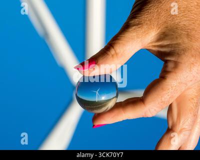 Gros plan d'une main avec des clous peints tenant une petite boule de verre qui reflète une éolienne contre un ciel bleu clair Banque D'Images