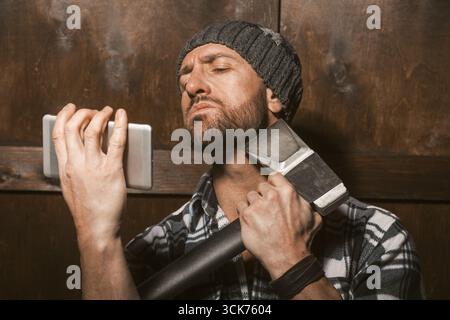 Un homme robuste, portant un bonnet et une chemise à carreaux, tient un smartphone d'une main tout en saisissant une hache de l'autre. Banque D'Images