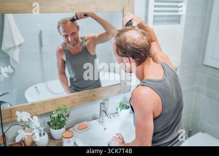 Concept corps et soin de la peau. Homme souriant se donnant une nouvelle coupe de cheveux à l'aide de tondeuse électrique rechargeable tout en regardant dans le miroir de salle de bain, Gro personnel Banque D'Images