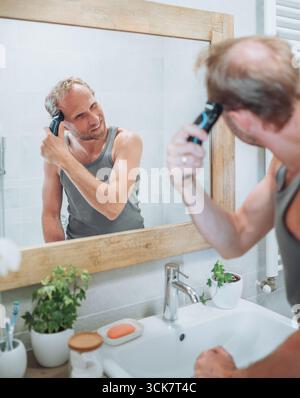 Concept corps et soin de la peau. Homme souriant se donnant une nouvelle coupe de cheveux à l'aide de tondeuse électrique rechargeable tout en regardant dans le miroir de salle de bain, Gro personnel Banque D'Images
