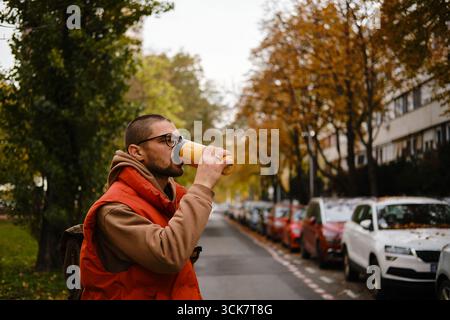 Jeune homme dans des verres buvant du thé chaud ou du café de thermos jaunes dans une rue de la ville avec des arbres d'automne et des voitures. Banque D'Images