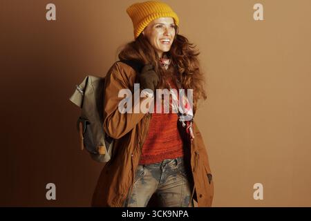 Une femme souriante avec de longs cheveux bruns bouclés, portant un bonnet jaune, une veste brune et un pull rouge, pose avec un sac à dos vert. Son pantalon camouflage fait allusion Banque D'Images