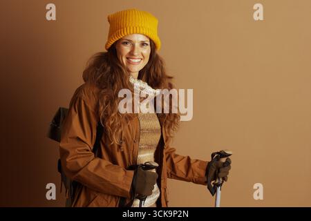 Une femme souriante avec de longs cheveux bruns bouclés, portant un chapeau tricoté jaune, une veste marron et un pull rayé, tient des bâtons de trekking et porte un dos Banque D'Images