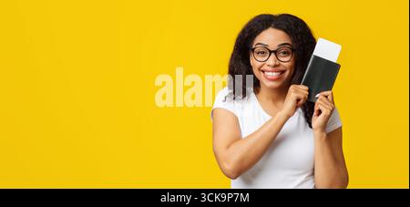 Touriste afro-américaine excitée avec passeport et billets d'avion se préparant pour les vacances, fond jaune Banque D'Images