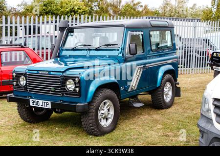Un Land Rover Defender bleu stationné sur l'herbe avec un fond d'arbres et de clôtures industrielles dans un cadre extérieur Banque D'Images