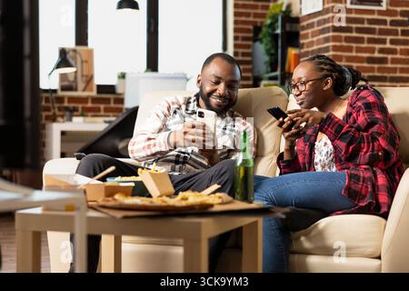 Homme barbu montrant joyeusement une vidéo drôle sur son appareil mobile à sa petite amie, assise confortablement sur le canapé. Couple afro-américain se liant dans le salon avec smartphones et nourriture à emporter. Banque D'Images