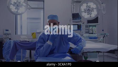 Chirurgien assis portant une tenue chirurgicale observant des moniteurs dans la salle d'opération, avec des plafonniers Banque D'Images